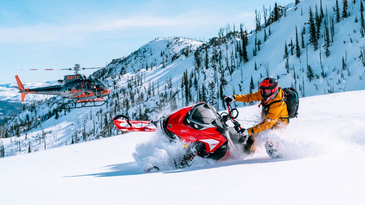 Ross Robinson riding a Lynx snowmobile with an helicopter in the bakcground