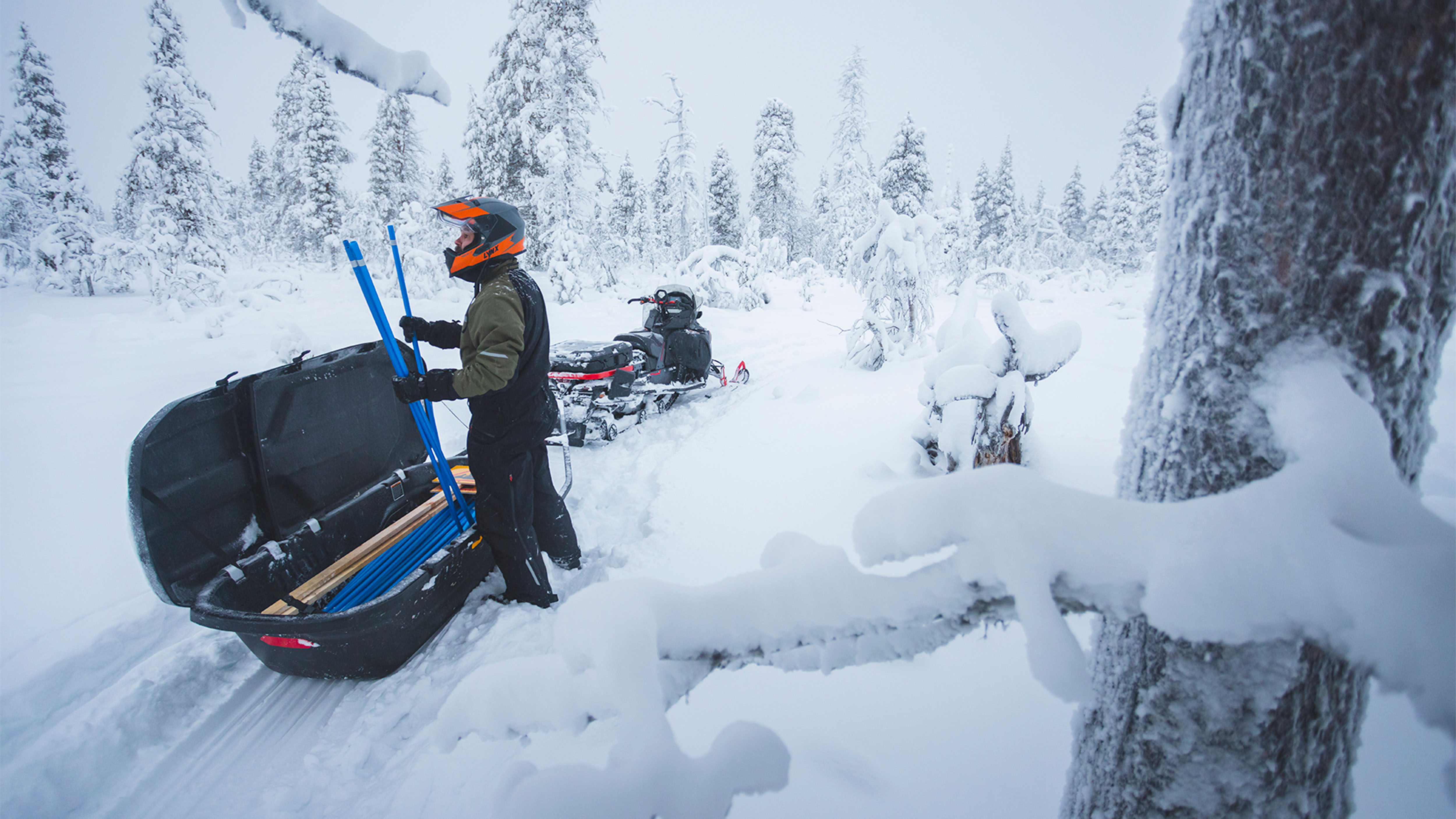 Mann tar skiltpinner bak på en Lynx Commander Limited-snøscooter i en snødekt skogsløype.