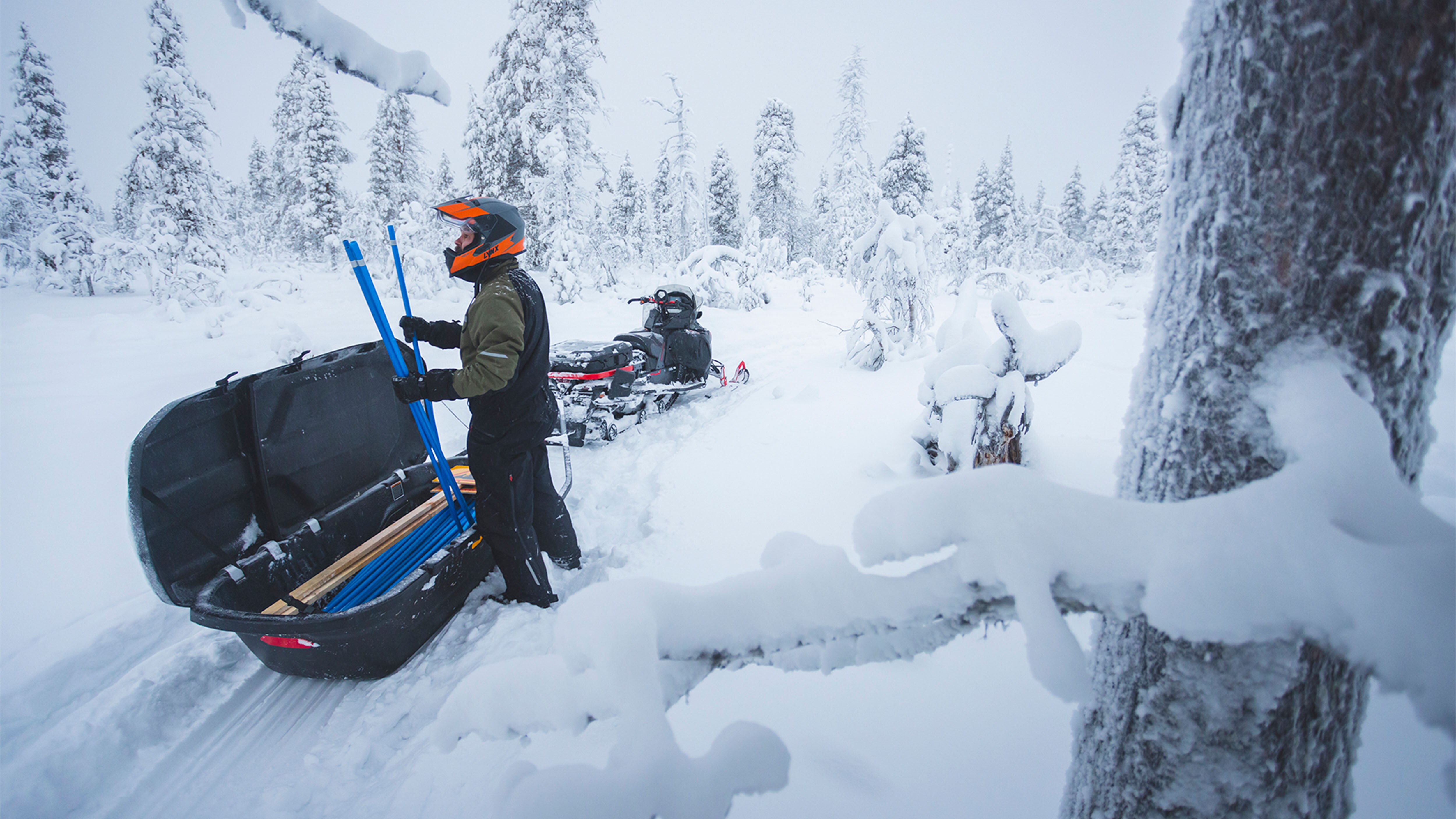 Mies ottamassa merkkitikkuja Lynx Commander Limited -moottorikelkan takana olevasta reestä lumisella metsäreitillä