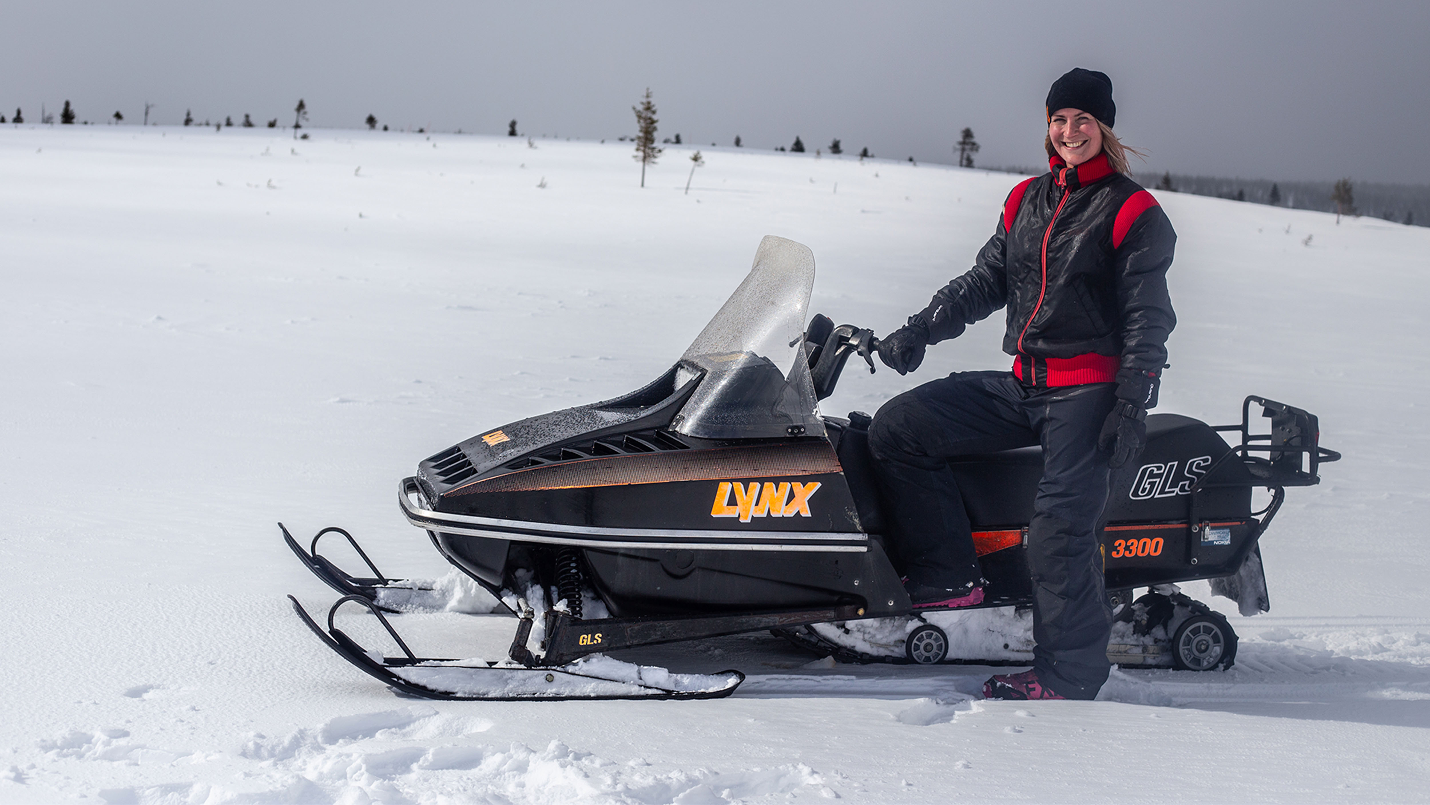 Leende kvinna står bredvid Lynx GLS 3300 snöskoter i fjällen