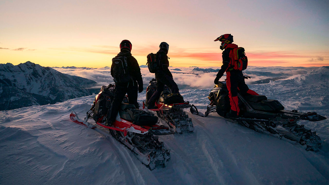 Trois motoneigistes sur leur Lynx Shredder au sommet d'une montagne pendant le coucher de soleil