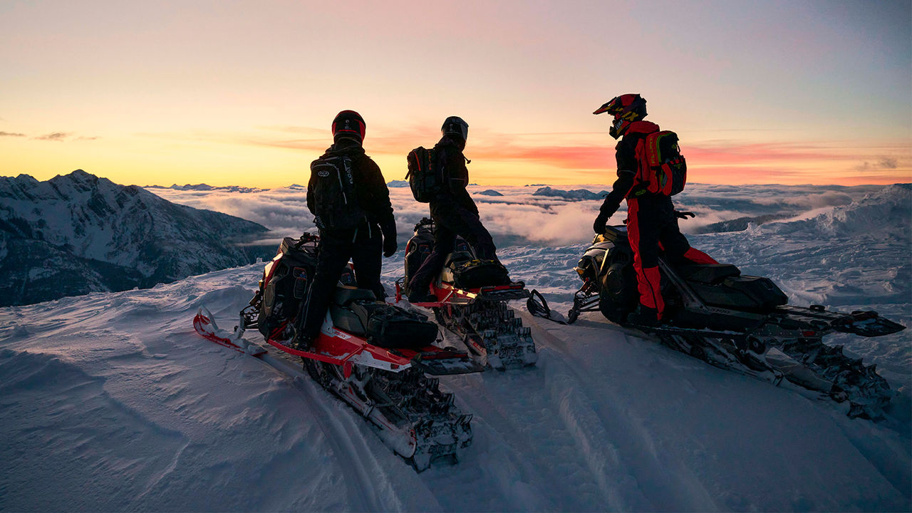 Kolme Lynx Shredder moottorikelkkaa pysäköitynä vuoren huipulle auringonlaskussa