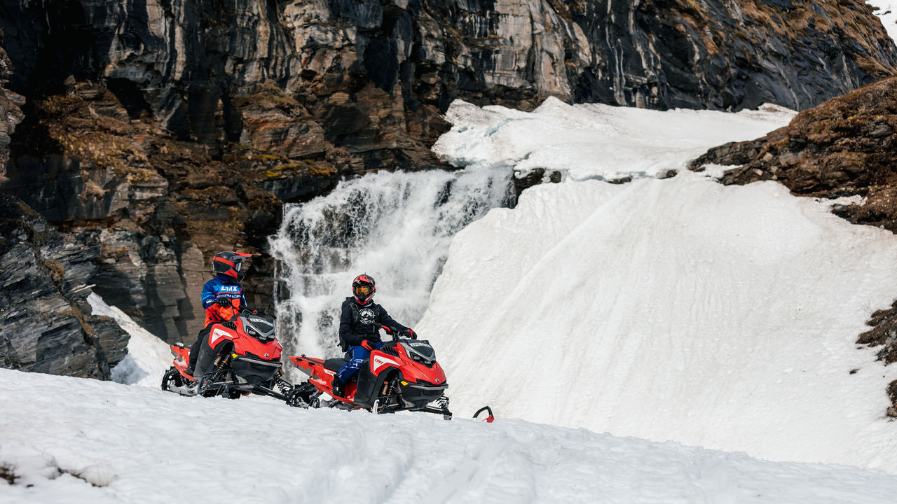 Two Lynx riders on their snowmobile next to a waterfall