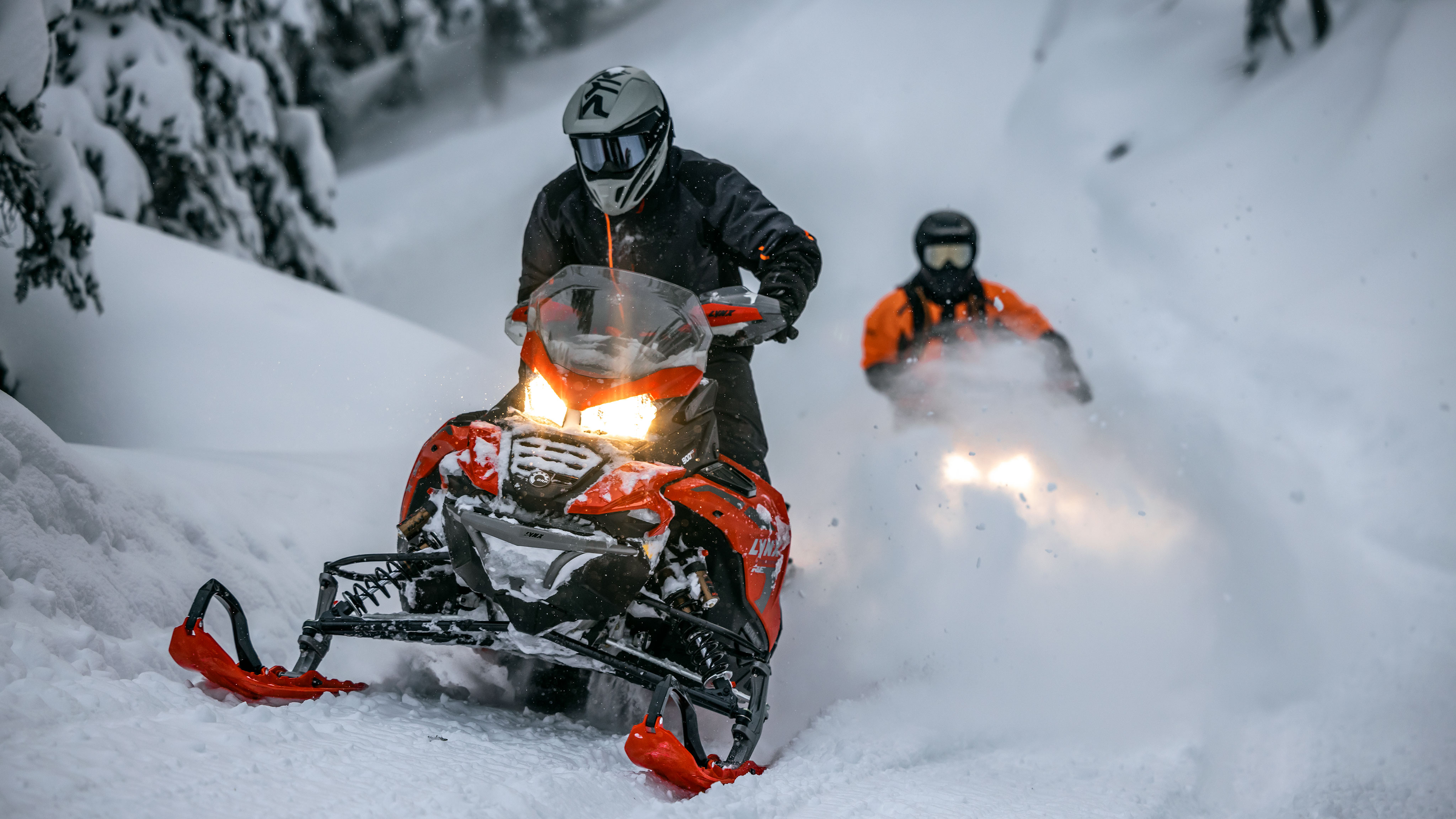 To førere som kjører Lynx-snøscooter gjennom en snødekt skog.