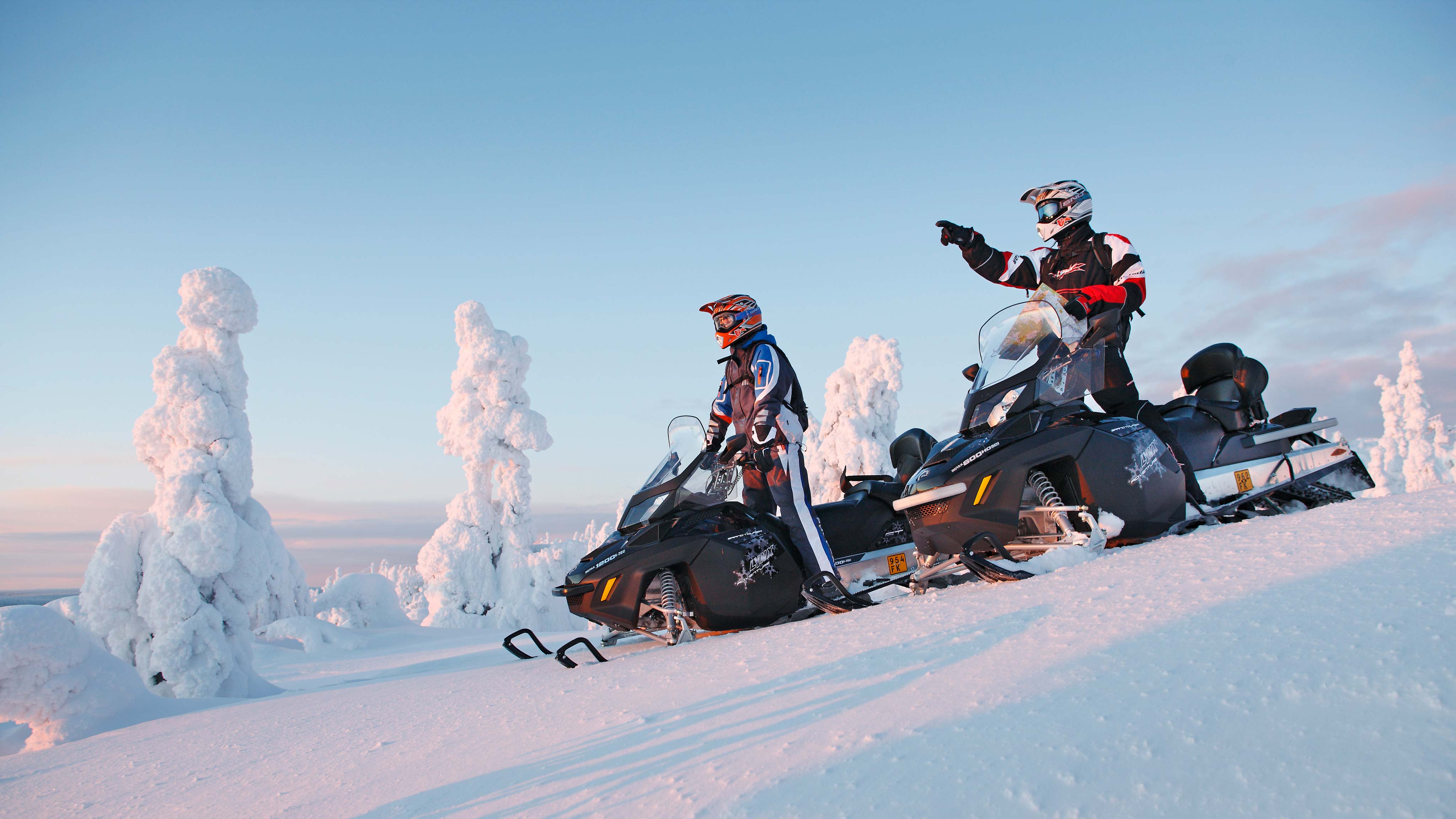 Men standing on two Lynx Adventure Grand Tourer snowmobiles and looking at the scenery