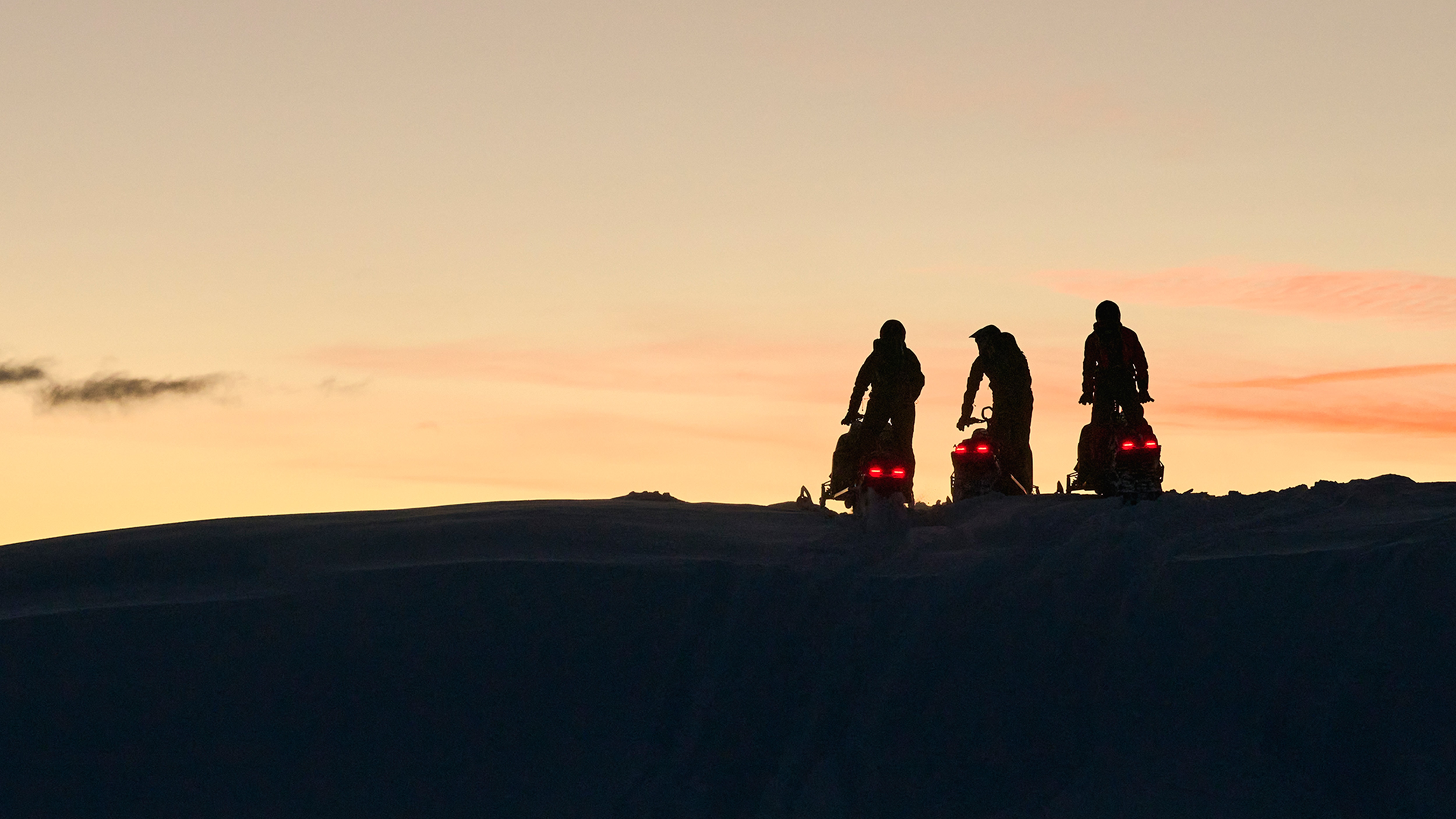 Three snowmobiles parked on top of hill at sunset