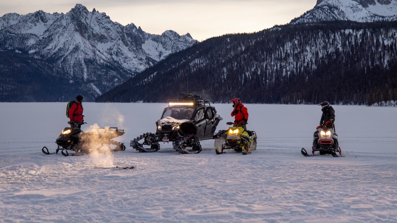 Quatre personnes en motoneiges Lynx et un véhicule tout‑terrain se rassemblent sur un paysage enneigé et gelé au crépuscule, entourés de montagnes alpines, pour une aventure hivernale en plein air.