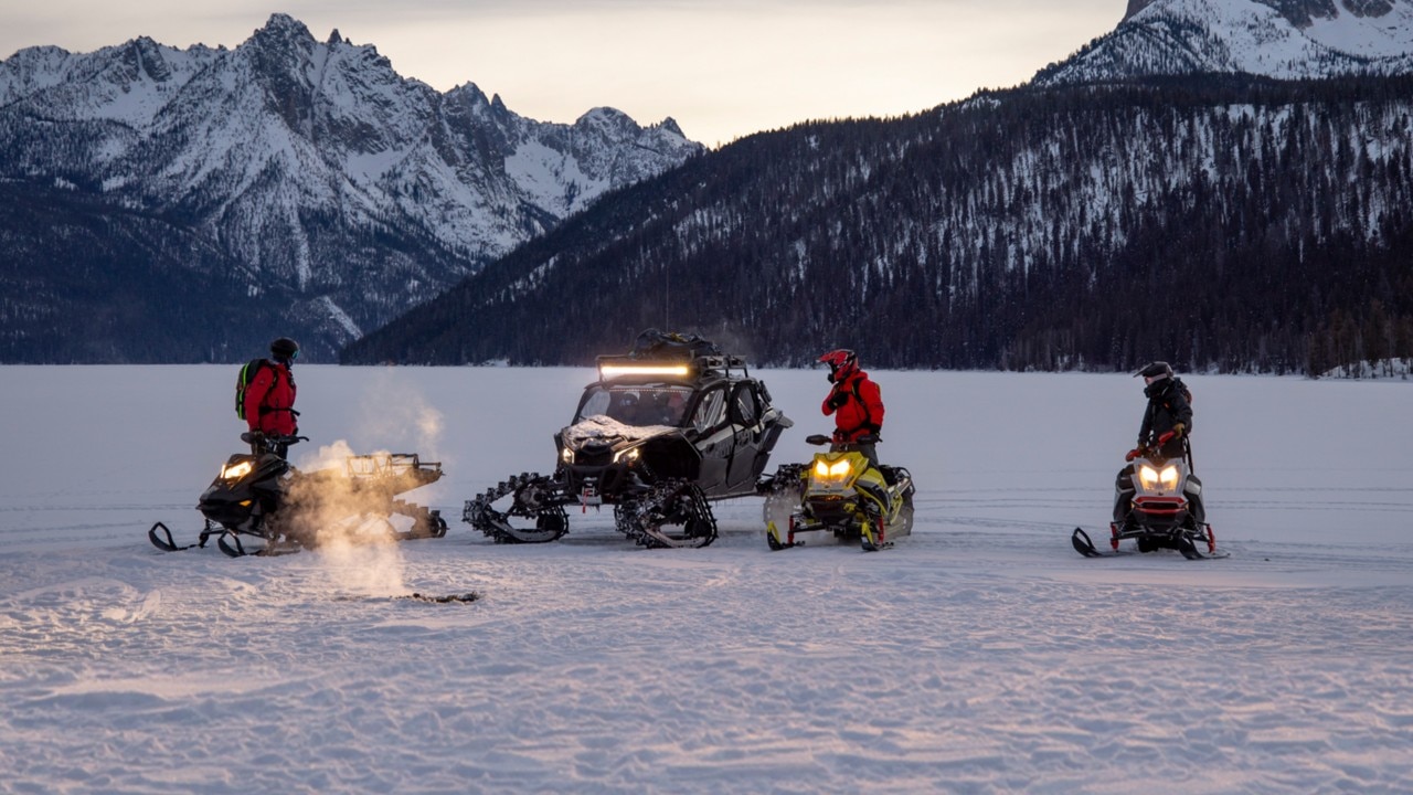 Four people on snowmobiles and an off-road vehicle gather on a snowy, frozen landscape at dusk, with mountains in the background